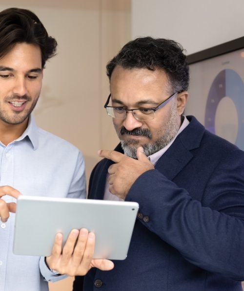 Positive Latin businessman showing project plan to mature Indian colleague Positive Latin businessman showing project plan on tablet to mature Indian colleague, consulting elder coworkers. Different aged diverse businessmen working with digital gadget together and talking